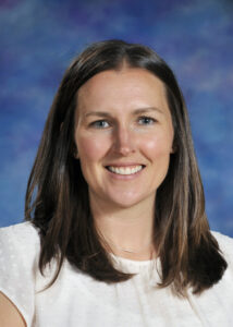 Portrait of a woman with long brown hair, wearing a white blouse, smiling against a blue background.