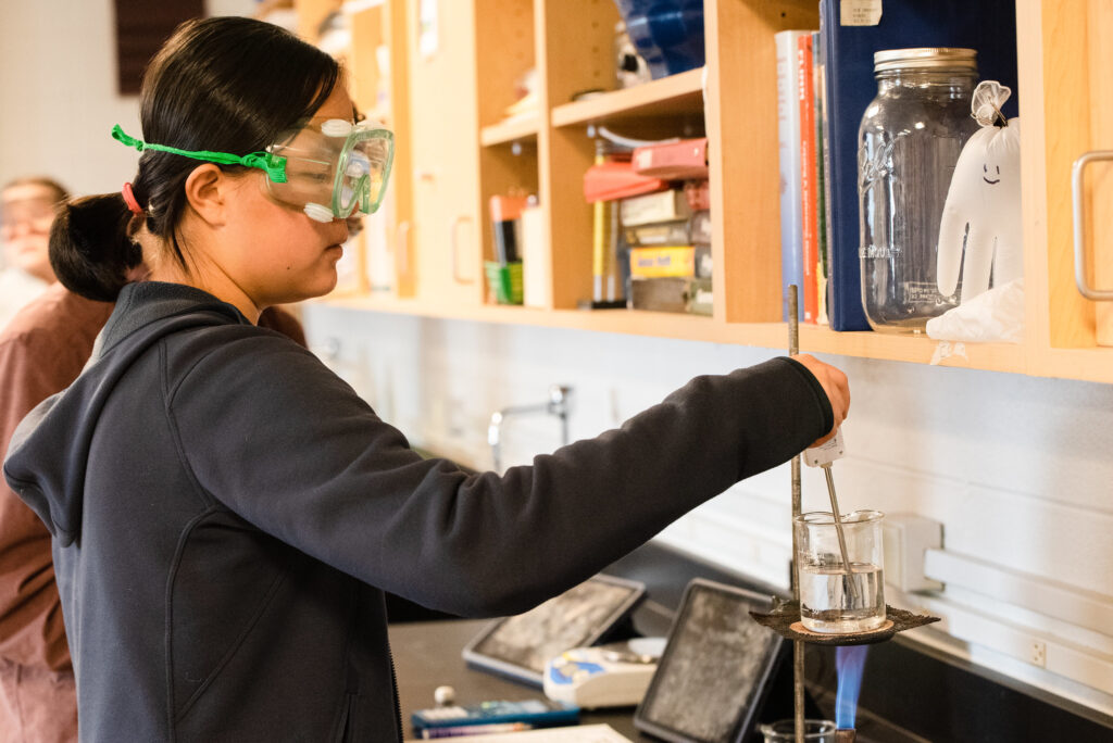 High school female in a classroom, wearing goggles while doing a science experiment. She's holding a thermometer in a beaker filled with boiling water. 