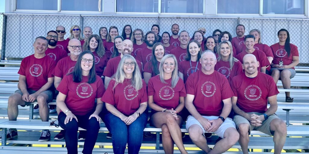 Group photo of Lyons Middle Senior High School staff. They are sitting on five rows of the bleachers outside by the football field and are facing the camera and smiling.
