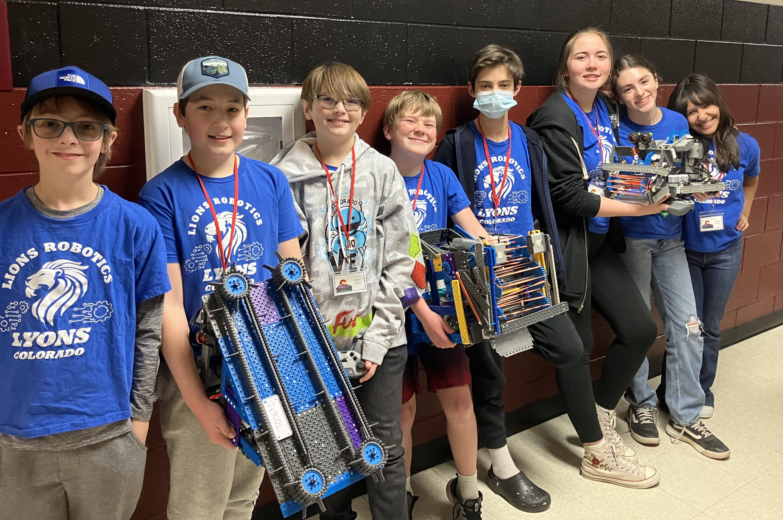 Standing in a line are five middle school boys on the left and three middle school girls on the right. They're holding robotics robots and trophies while smiling at the camera.