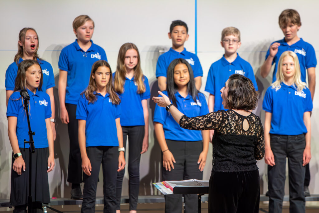 Two rows of middle school boys and girls standing on risers on a stage singing for audience. Female music teacher standing in front of them with her back to audience as she conducts the choir. 