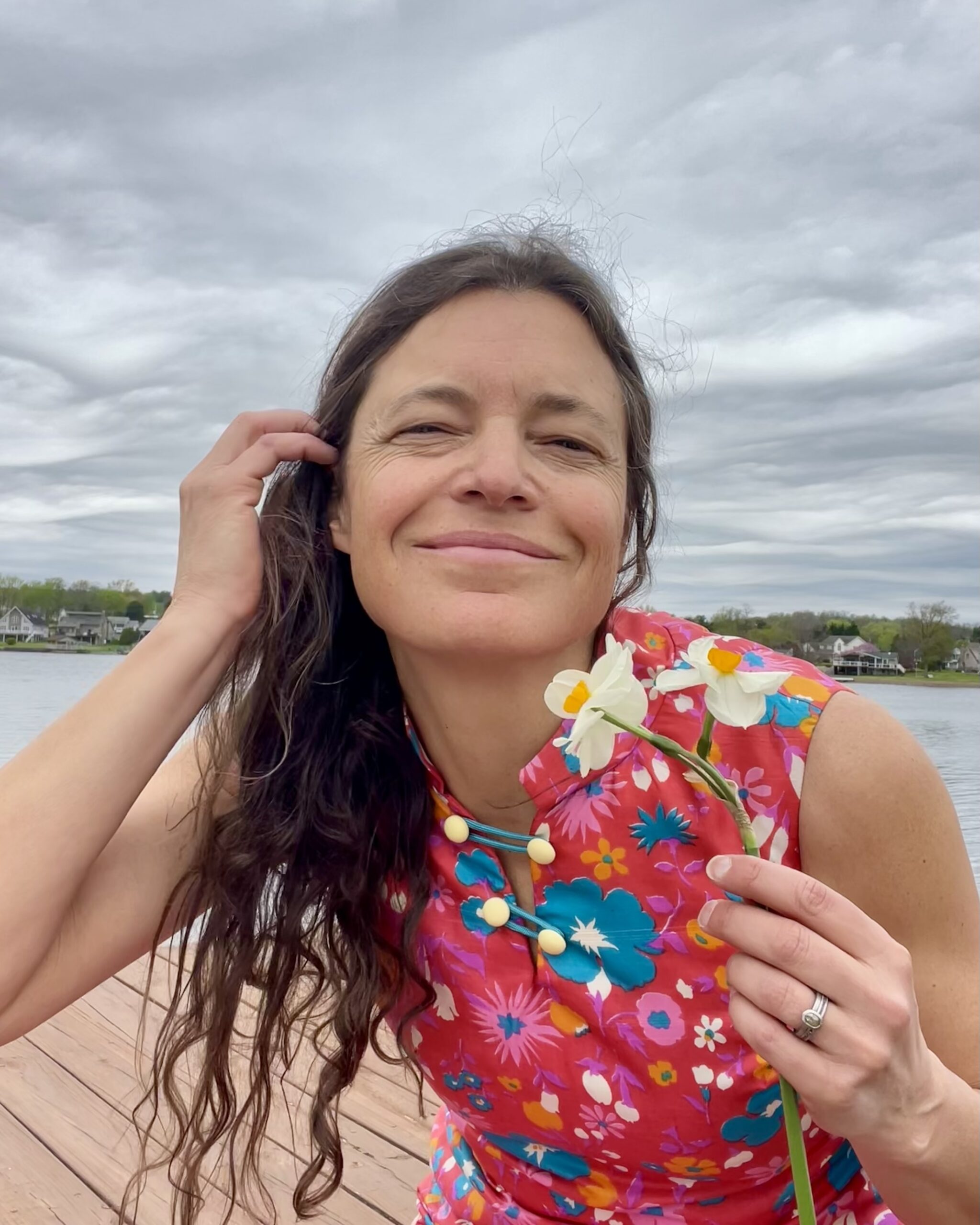 A woman with long hair smiles while holding a flower, wearing a colorful floral dress near a calm lake.