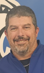 Smiling man with a beard and short hair, wearing a blue jacket against a blue and white background.