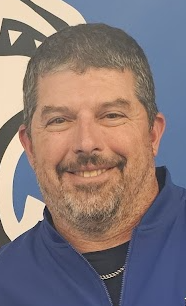 Smiling man with a beard and short hair, wearing a blue jacket against a blue and white background.