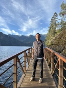 A person stands on a dock by a lake, surrounded by mountains and trees under a blue sky with clouds.
