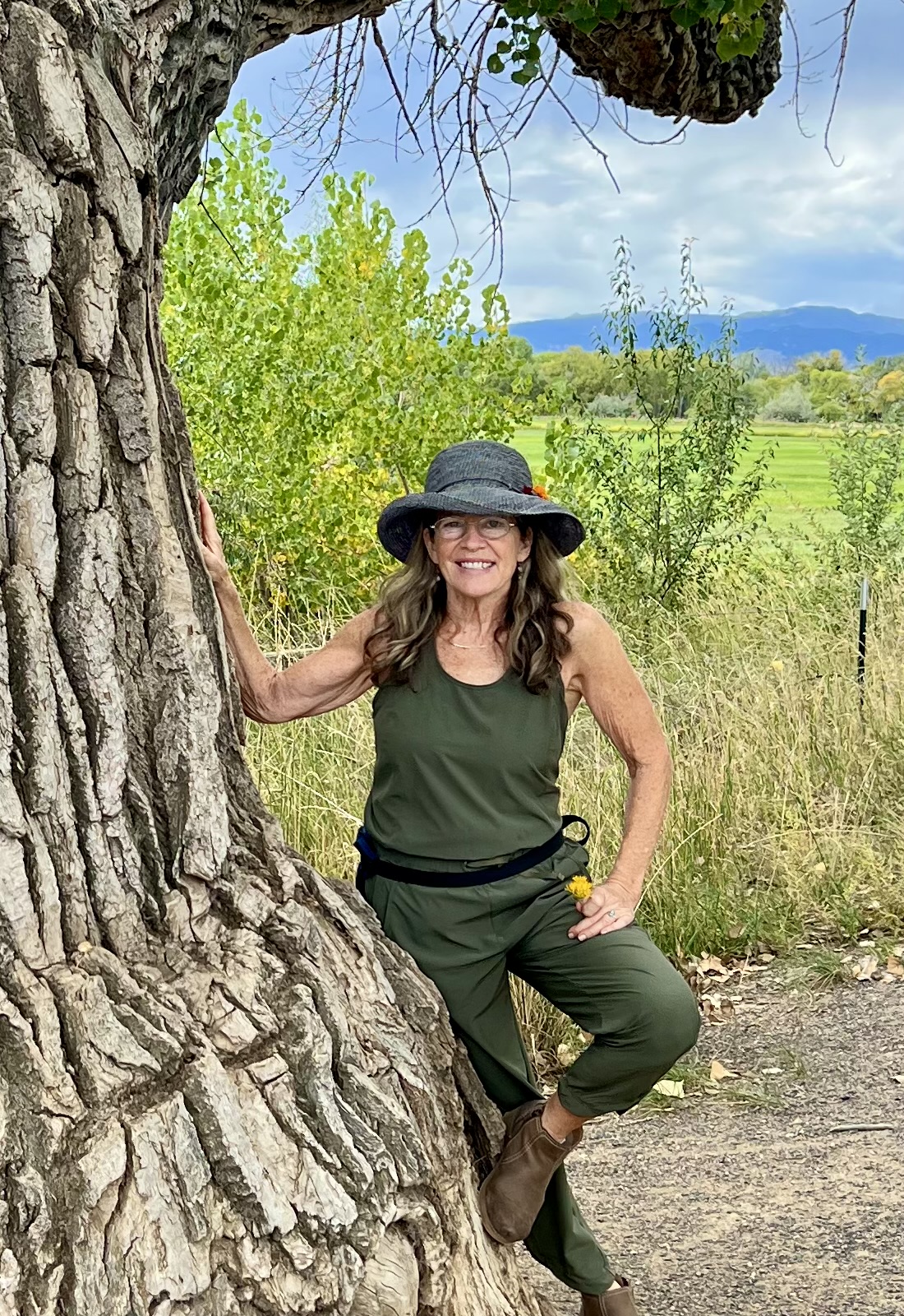 A woman in green clothing and a hat poses beside a large tree, with a scenic landscape of grass and trees in the background.
