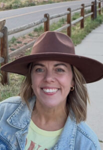 A smiling woman with short hair wears a wide-brimmed brown hat and denim jacket, standing on a path near a fence.