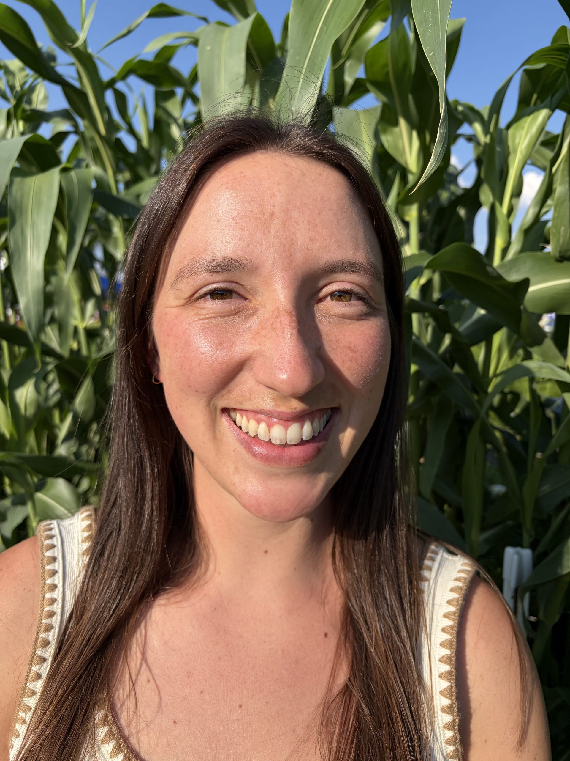A smiling woman with long brown hair stands in front of tall green corn plants under a clear blue sky.