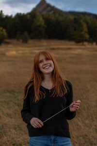 A smiling woman with long red hair stands in a grassy field, holding a small metal rod, with mountains behind her.