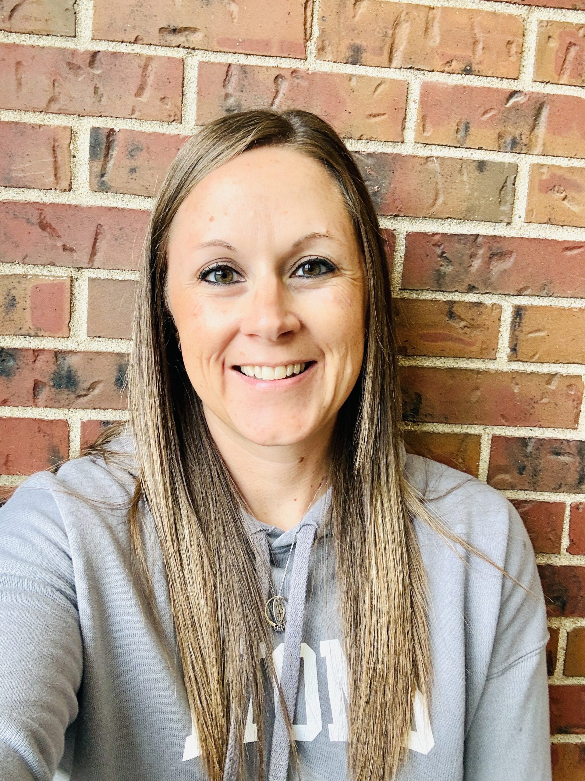 Smiling woman with long hair wearing a gray hoodie, posing against a brick wall background.