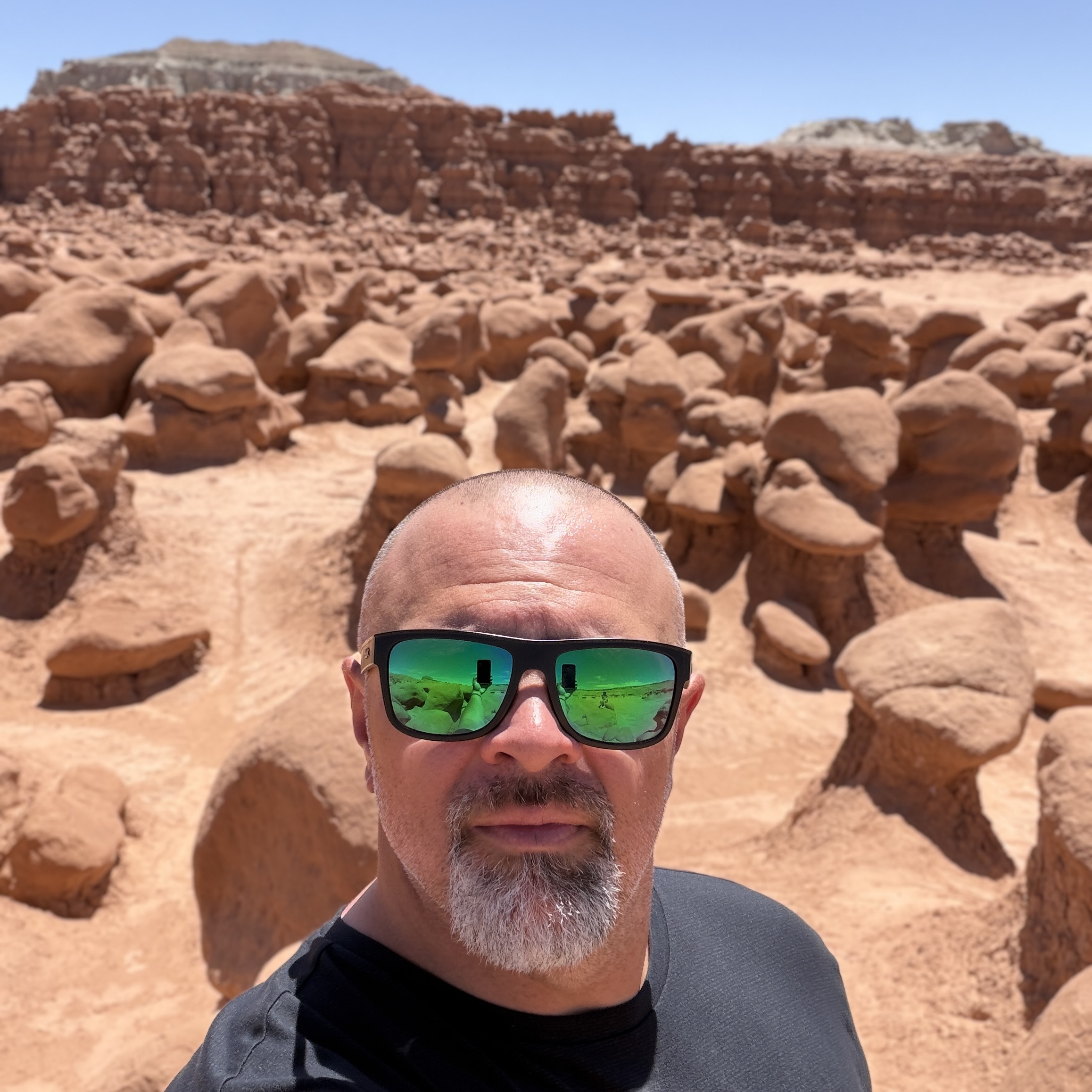 Una persona con gafas de sol posa en un paisaje desértico repleto de formaciones rocosas singulares, bajo un cielo azul y despejado.
